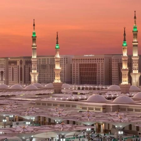 다르 알 이만 인터컨티넨탈 호텔 메디나 외부 사진 The photo depicts a scenic view of a mosque complex, likely during sunset, with several minarets illuminated and showcasing green lights. The architecture features domed roofs, and the setting includes a backdrop of buildings, all bathed in a warm, o