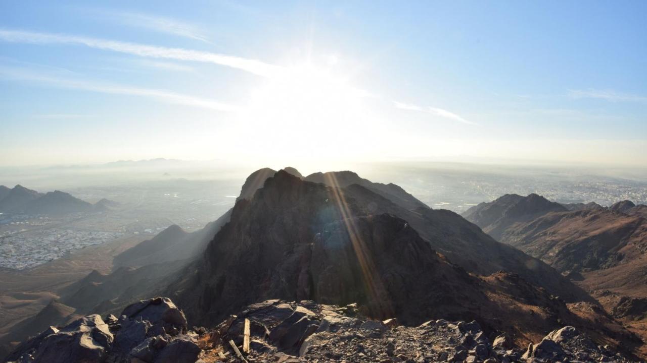다르 알 이만 인터컨티넨탈 호텔 메디나 외부 사진 The photo shows a scenic view from a mountain top, with peaks silhouetted against the bright sun. The sun is positioned low in the sky, creating a warm glow and illuminating the landscape below. In the foreground, there are rocky terrains, while the 
