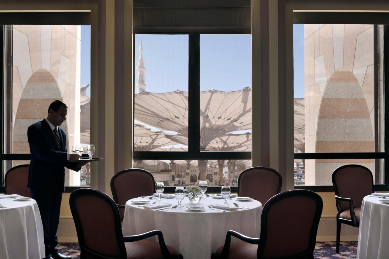 다르 알 이만 인터컨티넨탈 호텔 메디나 외부 사진 The photo shows a dining area with elegant decor. A waiter is standing to the side, holding a tray. The tables are neatly set with white tablecloths and fine dining arrangements. Large windows provide a view of an outdoor area featuring architectural