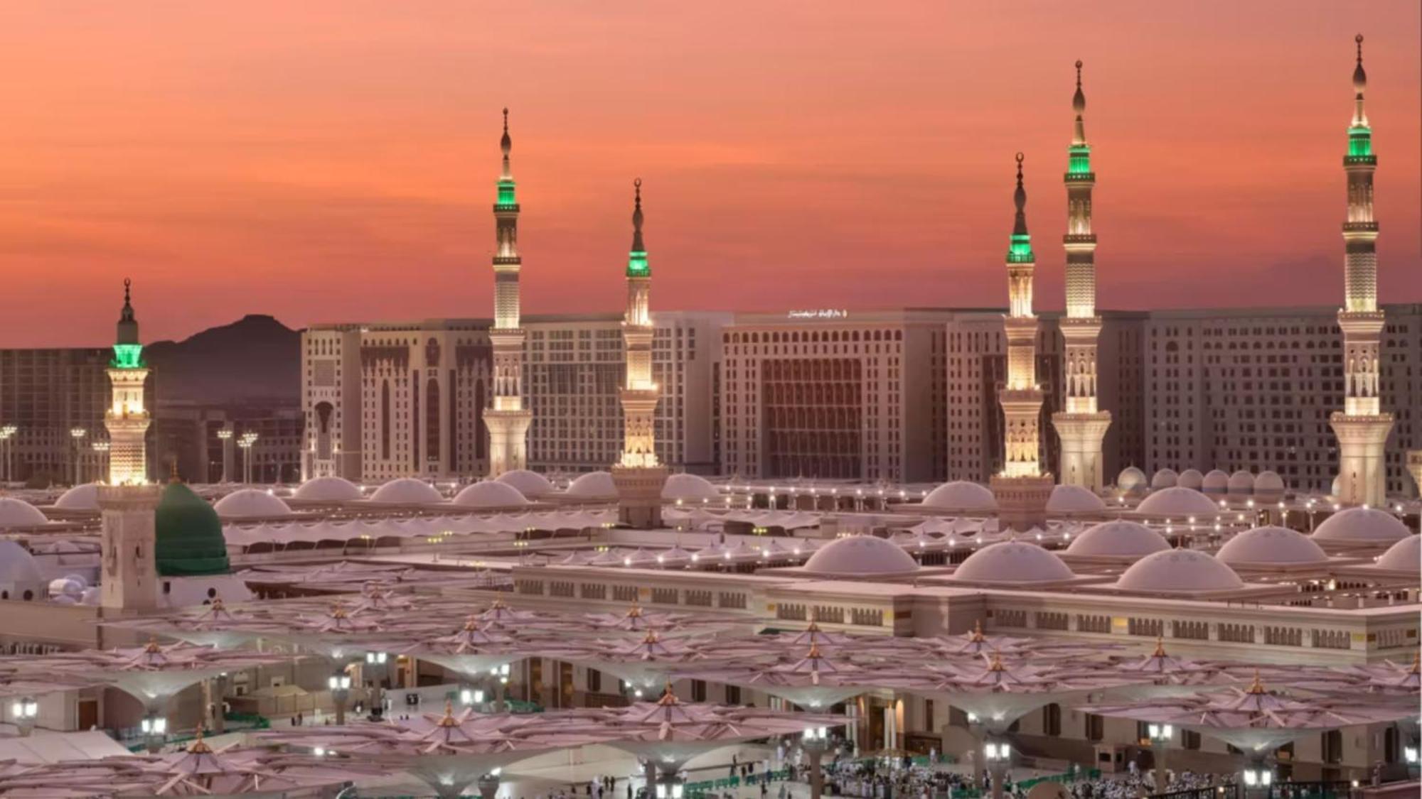 다르 알 이만 인터컨티넨탈 호텔 메디나 외부 사진 The photo depicts a scenic view of a mosque complex, likely during sunset, with several minarets illuminated and showcasing green lights. The architecture features domed roofs, and the setting includes a backdrop of buildings, all bathed in a warm, o