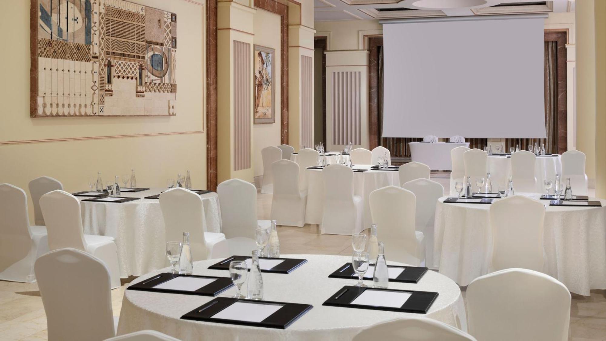 다르 알 이만 인터컨티넨탈 호텔 메디나 외부 사진 The photo depicts an elegantly arranged event space. There are several round tables covered with white tablecloths and surrounded by white chairs. Each table is set with a black folder or menu and a glass of water. The walls are adorned with artworks