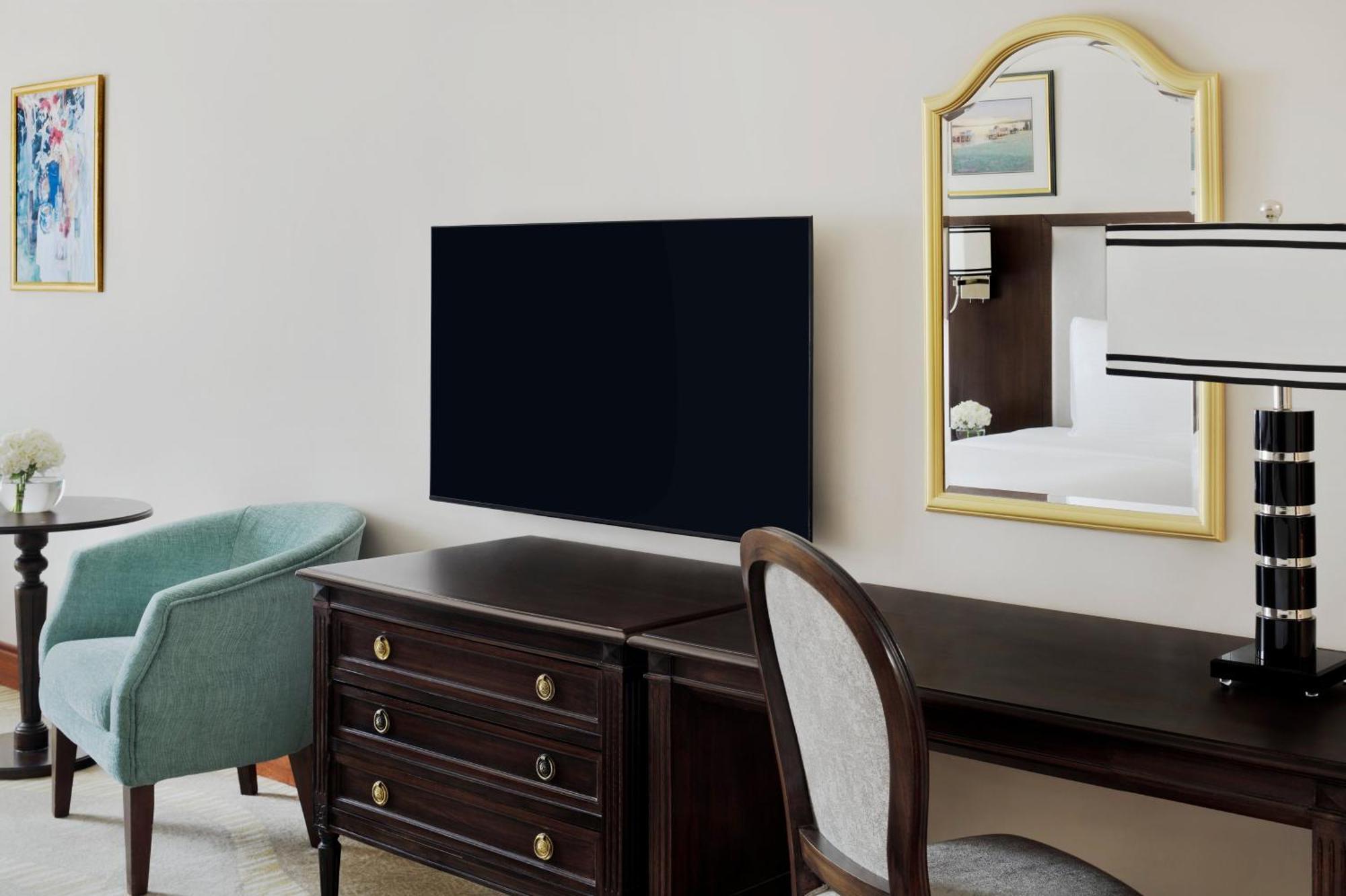 다르 알 이만 인터컨티넨탈 호텔 메디나 외부 사진 The photo shows a hotel room interior featuring a mounted flat-screen television on the wall. Below the TV, there is a dark wooden dresser with drawers. To the right, there's a wooden desk and a chair with a light-colored seat. A small table with a l