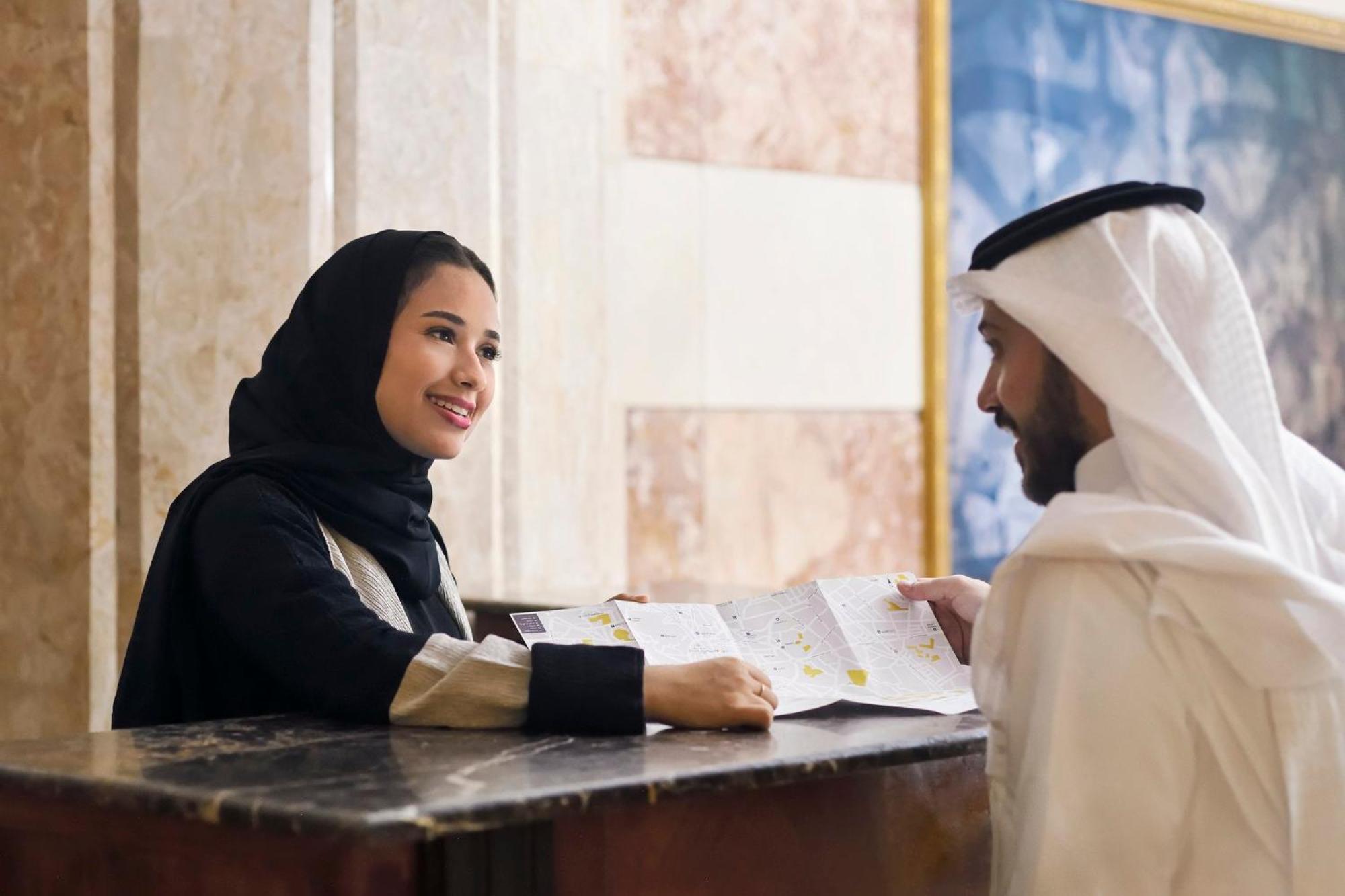 다르 알 이만 인터컨티넨탈 호텔 메디나 외부 사진 The photo depicts a woman in a black hijab and a light-colored outfit smiling as she interacts with a man wearing traditional Emirati attire, including a white kandura and ghutrah (headscarf). They are at a reception desk in what appears to be a hote
