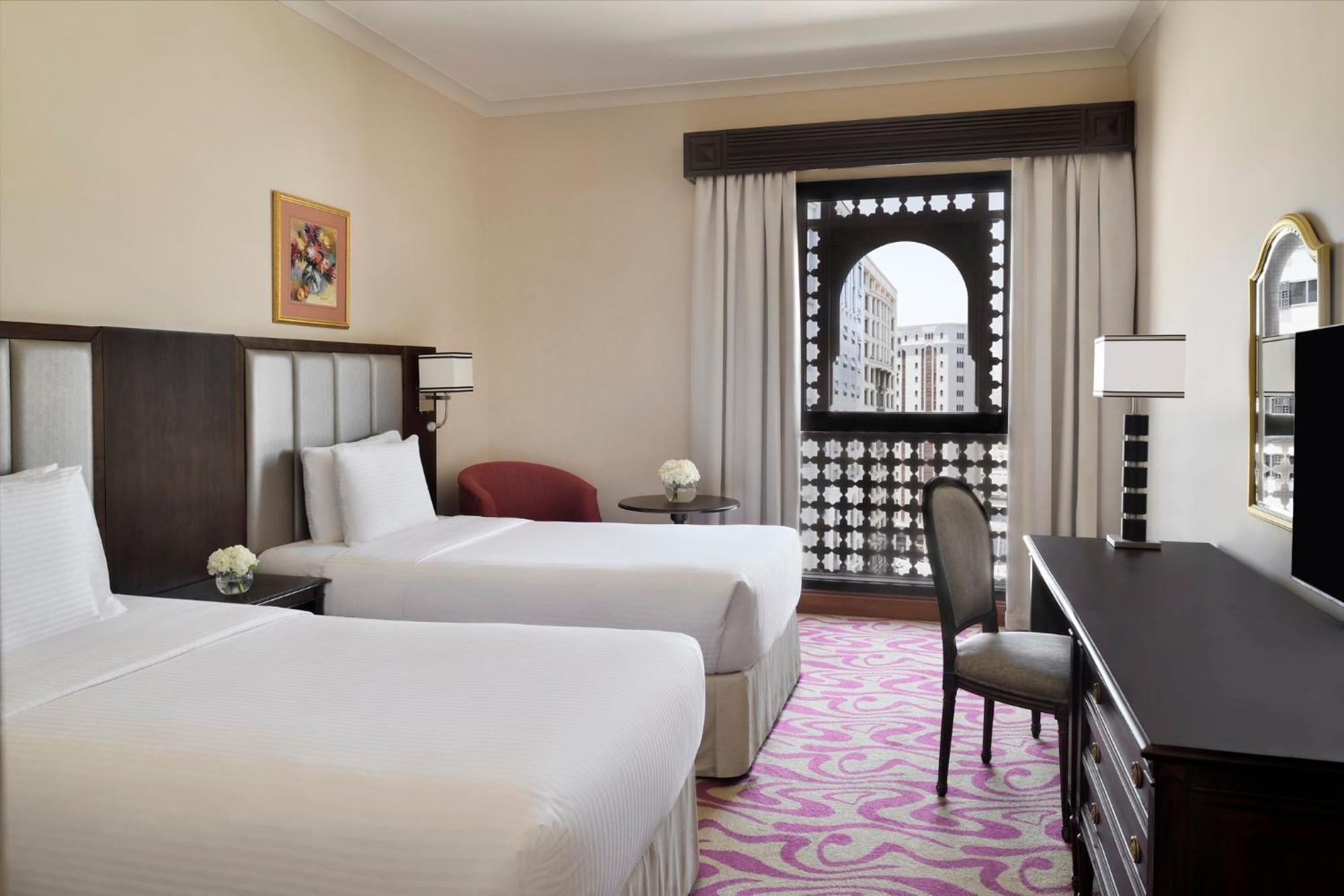 다르 알 이만 인터컨티넨탈 호텔 메디나 외부 사진 The photo shows a hotel room featuring two single beds with white bedding. The room has a light, neutral color scheme and is decorated with minimal furnishings. There's a small seating area with a red chair and a round table, as well as a desk with a