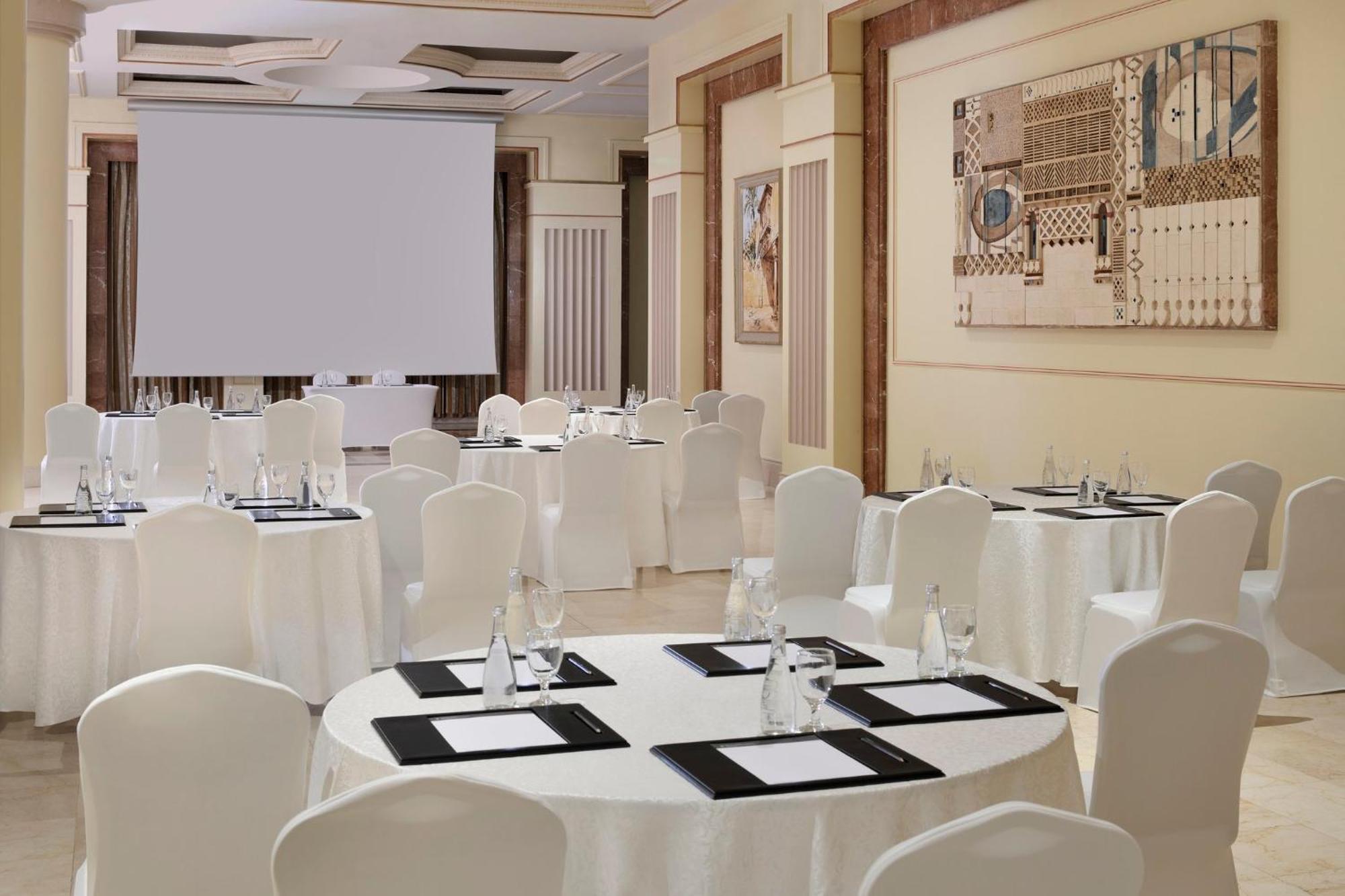 다르 알 이만 인터컨티넨탈 호텔 메디나 외부 사진 The photo shows a well-decorated event space or meeting room. It features several round tables covered with white tablecloths and surrounded by white chairs. Each table has black placemats and glass water bottles, suggesting a formal setup for a gath