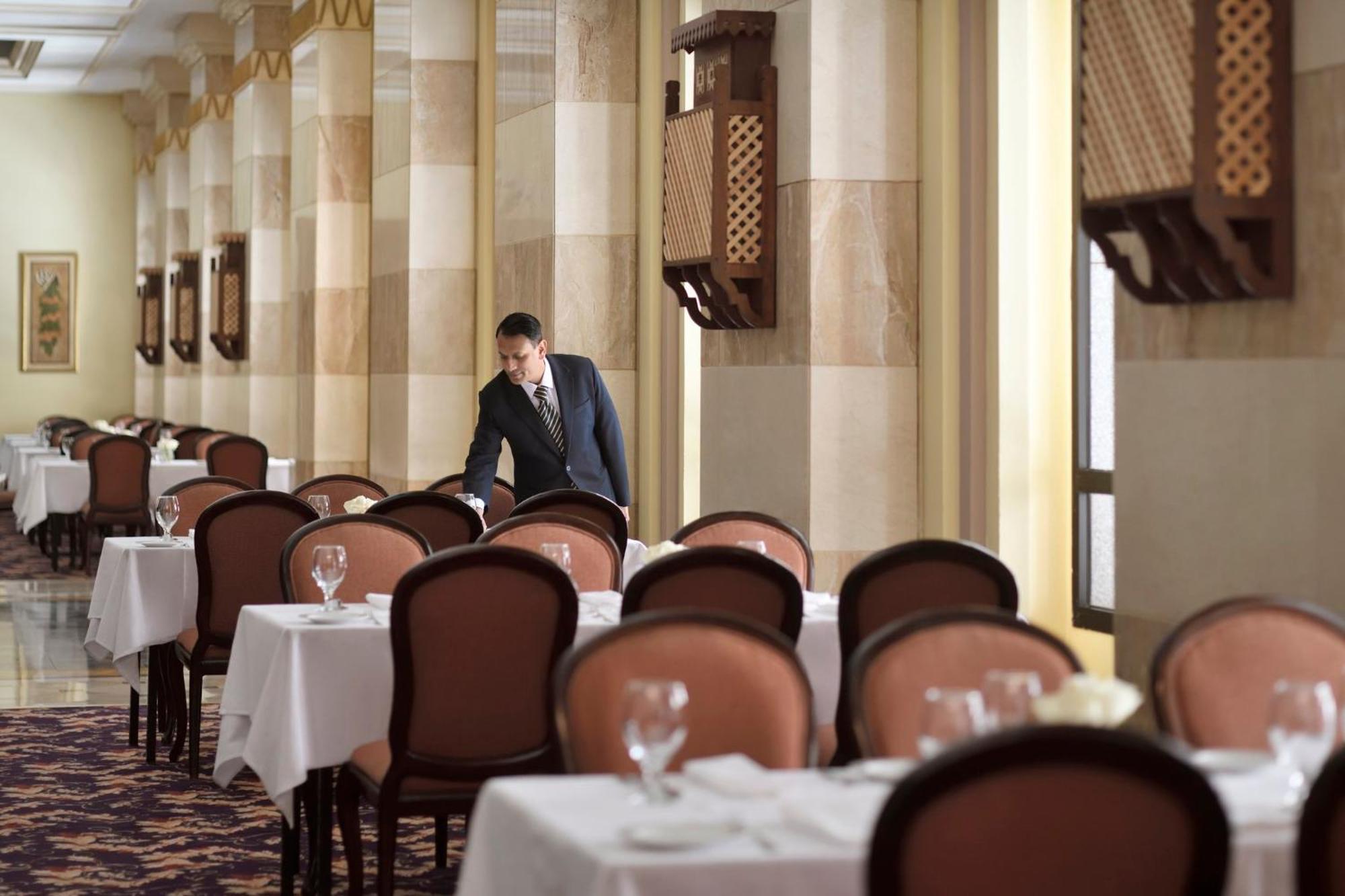 다르 알 이만 인터컨티넨탈 호텔 메디나 외부 사진 The photo depicts a dining area in an elegant restaurant. There are several tables set with white tablecloths, each accompanied by chairs. A waiter, dressed in a formal suit, is seen arranging or tending to one of the tables. The restaurant features 