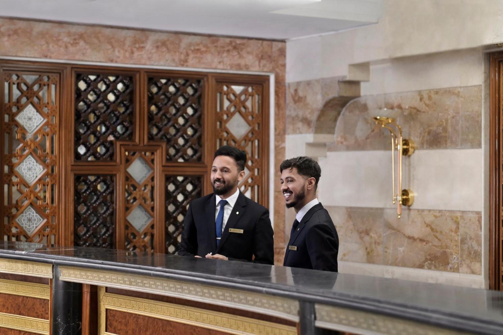 다르 알 이만 인터컨티넨탈 호텔 메디나 외부 사진 The photo shows two men standing behind a reception desk in an elegant setting, likely a hotel or similar establishment. They are both dressed in formal attire, wearing suits and ties, and appear to be smiling or engaging in conversation. The backgro