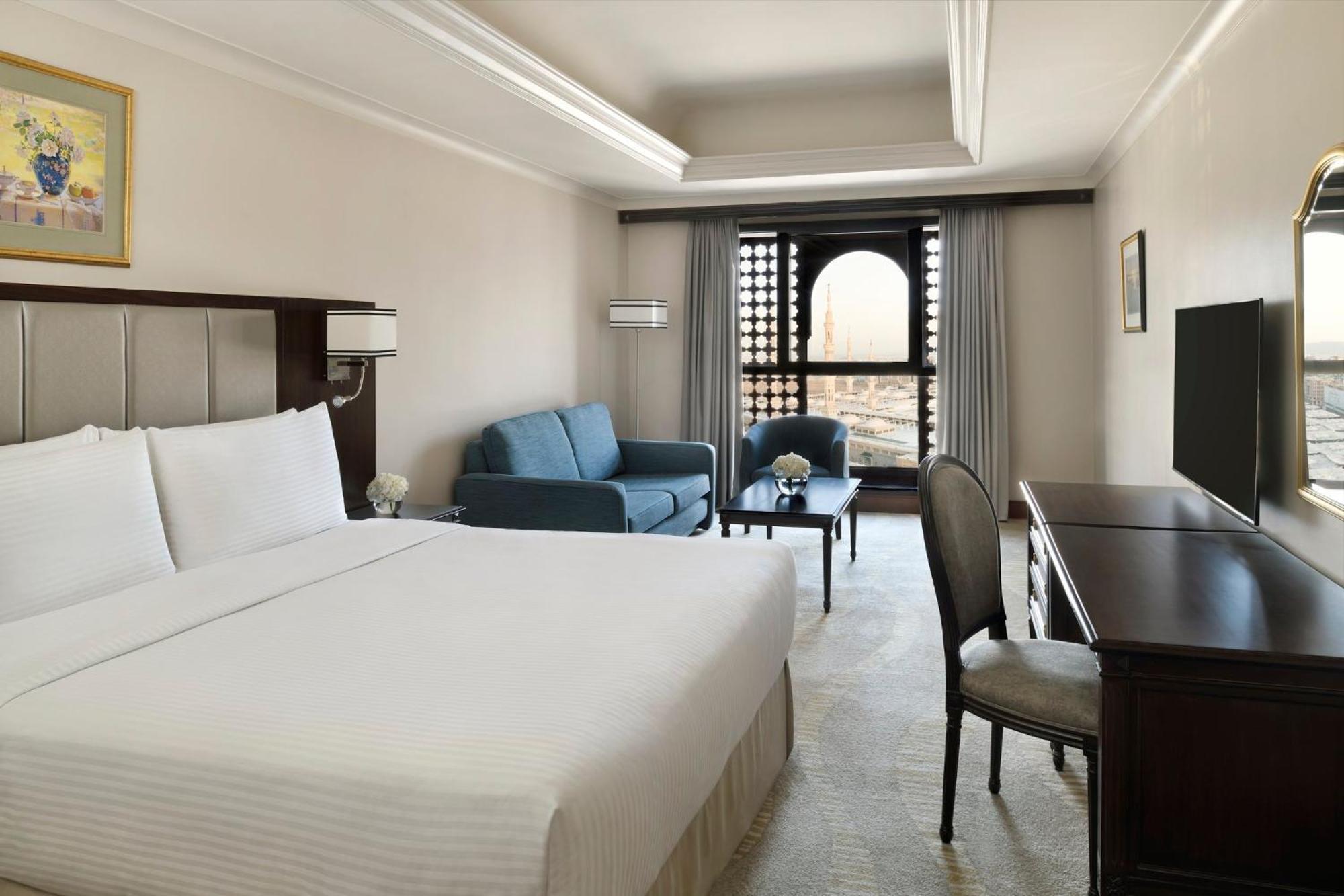 다르 알 이만 인터컨티넨탈 호텔 메디나 외부 사진 The photo shows a hotel room designed with a modern and comfortable aesthetic. It features a large bed with white linens, a seating area with a blue sofa, and a small table. There is a wooden desk with a chair and a television opposite the bed. Natur