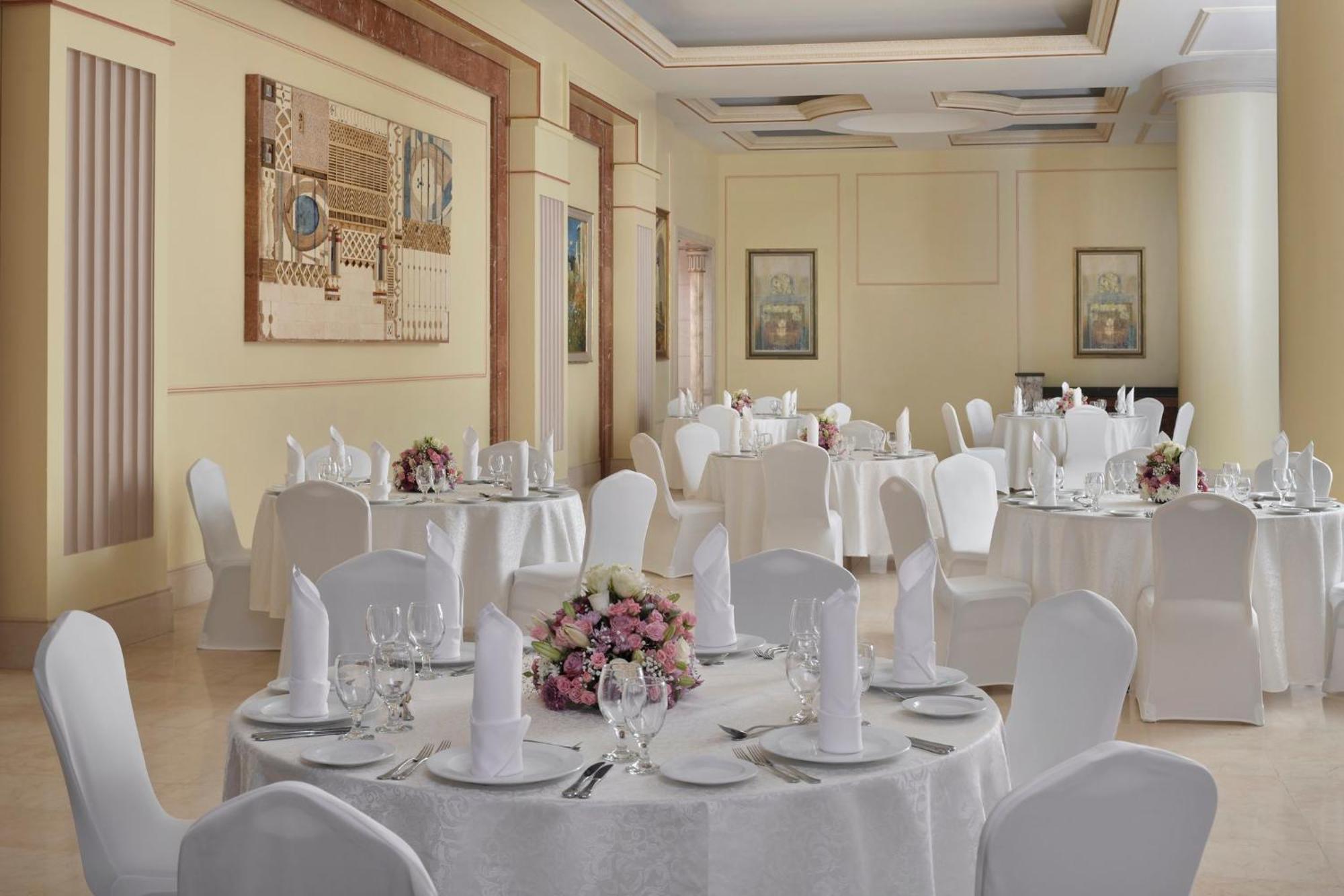다르 알 이만 인터컨티넨탈 호텔 메디나 외부 사진 The photo shows an elegantly arranged dining or function room. There are round tables covered with white tablecloths, each set with fine dining ware including glassware, utensils, and neatly folded white napkins. Some tables have floral centerpieces 