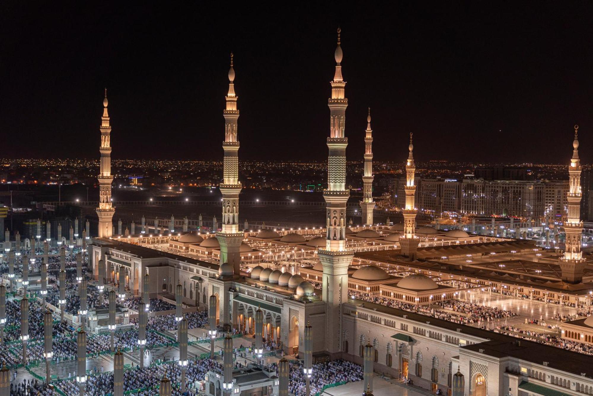 다르 알 이만 인터컨티넨탈 호텔 메디나 외부 사진 The photo depicts a stunning nighttime view of a large mosque complex, prominently featuring tall minarets that are illuminated against the dark sky. The surrounding area is bustling with people, likely worshippers, gathered in the courtyard of the m