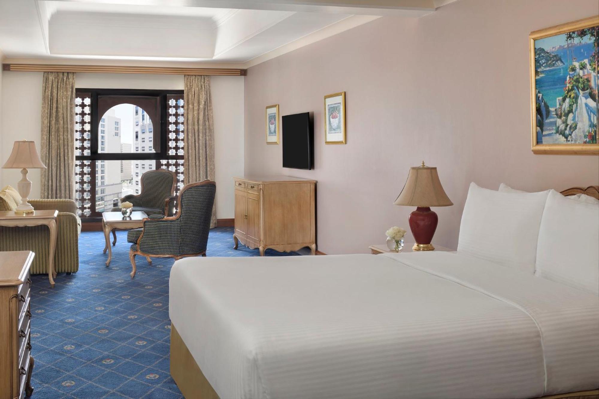 다르 알 이만 인터컨티넨탈 호텔 메디나 외부 사진 The photo shows a well-furnished hotel room. It features a large bed with a white duvet, two bedside lamps, and decorative paintings on the walls. There is a comfortable seating area with two chairs and a small table. A wooden dresser is positioned a