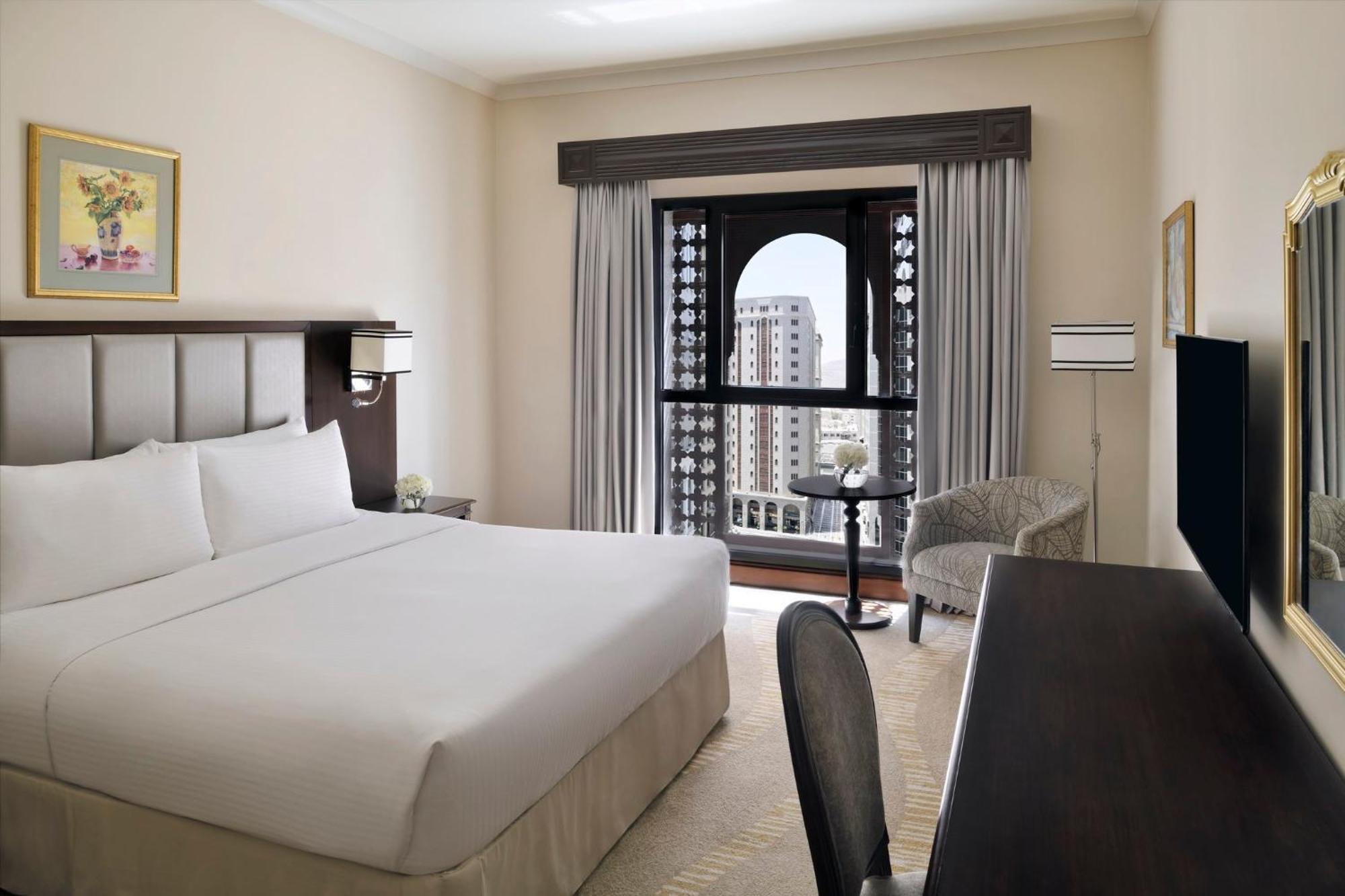 다르 알 이만 인터컨티넨탈 호텔 메디나 외부 사진 The photo shows a hotel room with a modern and minimalistic design. It features a large bed with white linens, a side table, and a comfortable chair near a window. The window has a view of a cityscape, and elegant curtains frame the window. There is 