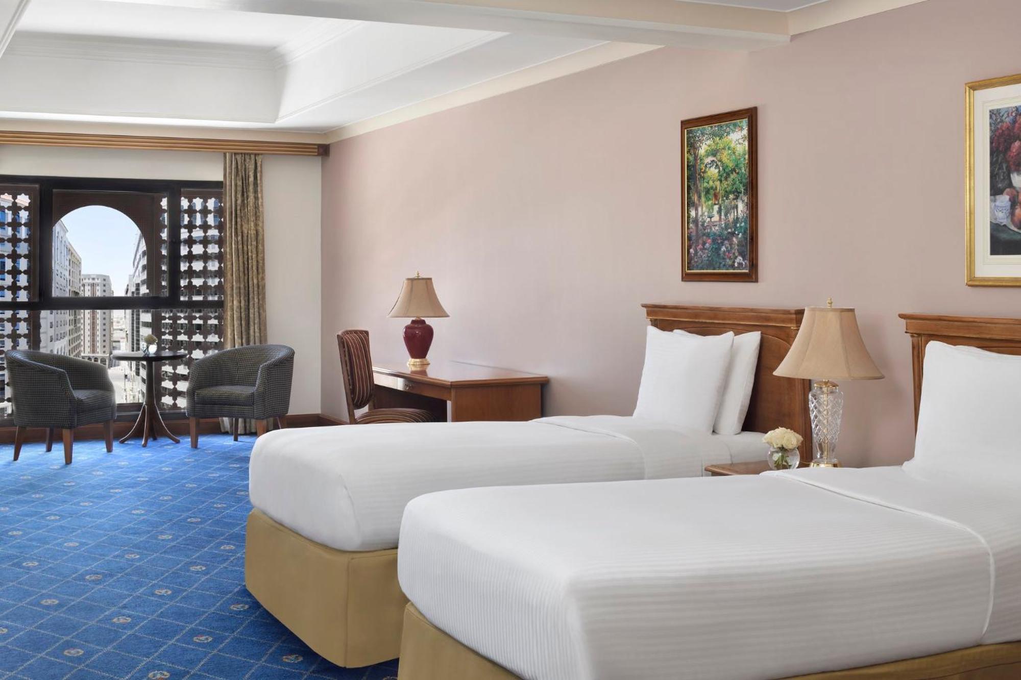 다르 알 이만 인터컨티넨탈 호텔 메디나 외부 사진 The photo shows a nicely designed hotel room featuring two twin beds with white linens and beige bases. In the background, there's a wooden desk and a lamp, along with a pair of comfortable armchairs positioned near a large window. The window showcas