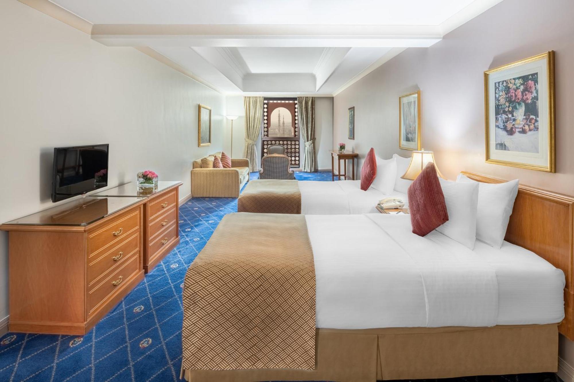 다르 알 이만 인터컨티넨탈 호텔 메디나 외부 사진 The photo shows a hotel room featuring two beds with white linens and decorative pillows. There is a wooden dresser with a television on it. The flooring has a blue carpet with a pattern, and the walls are painted in a light color. The room is well-l