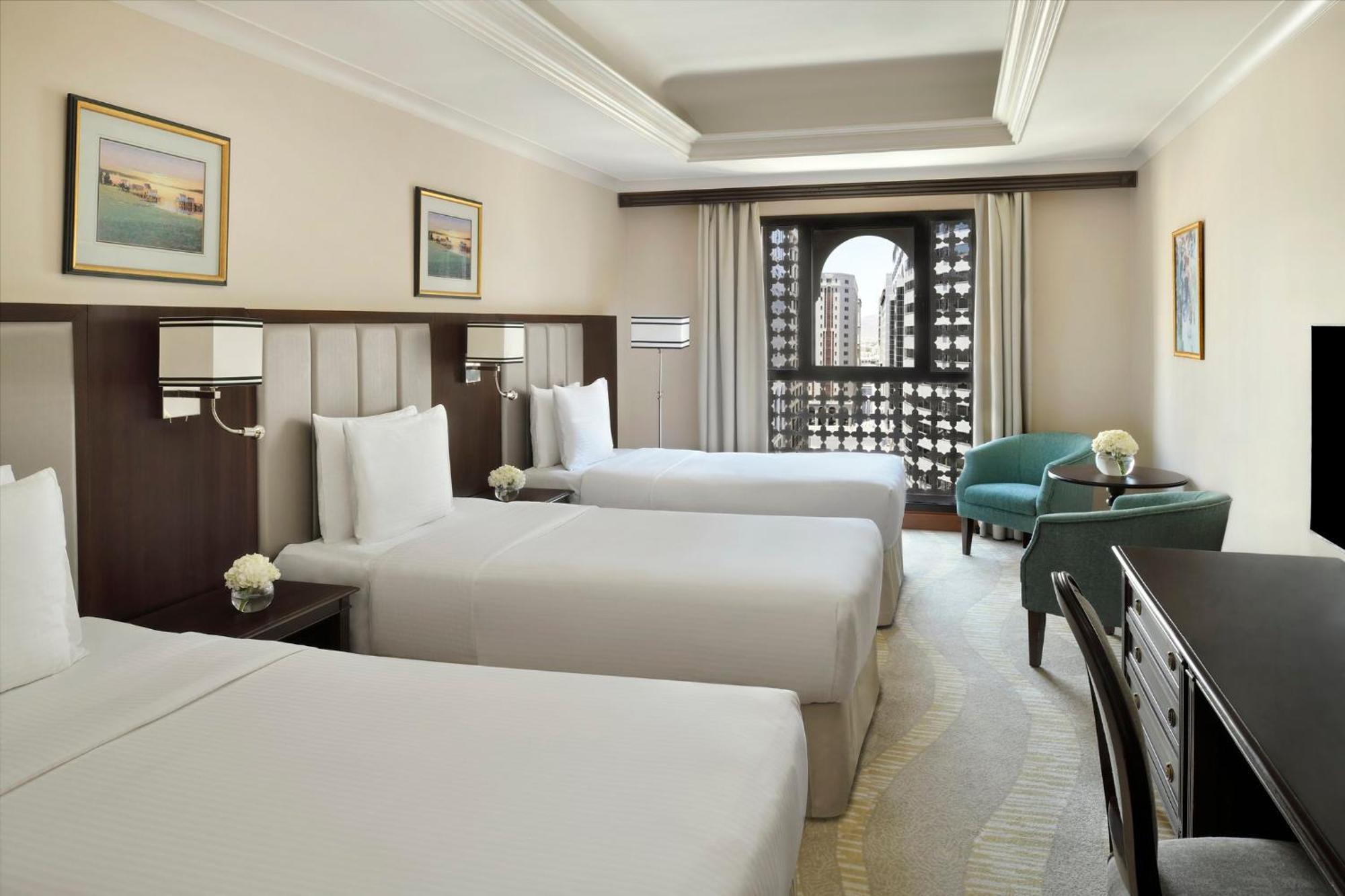 다르 알 이만 인터컨티넨탈 호텔 메디나 외부 사진 The photo shows a hotel room with three single beds, dressed in white linens. There are two paintings on the walls, depicting serene landscapes. A large window allows natural light to enter, and it is adorned with elegant curtains. A smaller seating 
