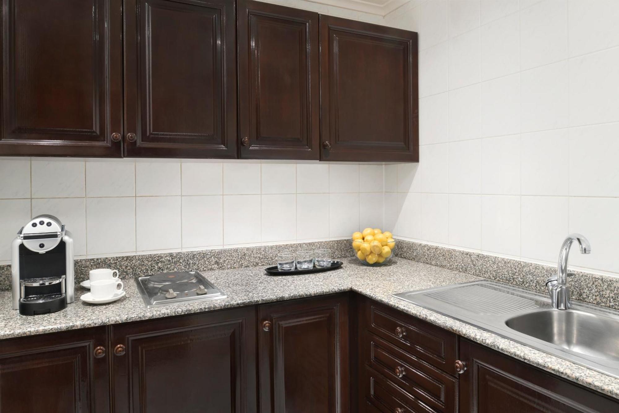 다르 알 이만 인터컨티넨탈 호텔 메디나 외부 사진 The photo shows a kitchen with dark wooden cabinetry and a light-colored countertop. There are a few appliances visible, including a coffee machine and a stovetop with burners. To the right, there is a sink, and a bowl of yellow lemons is placed on t