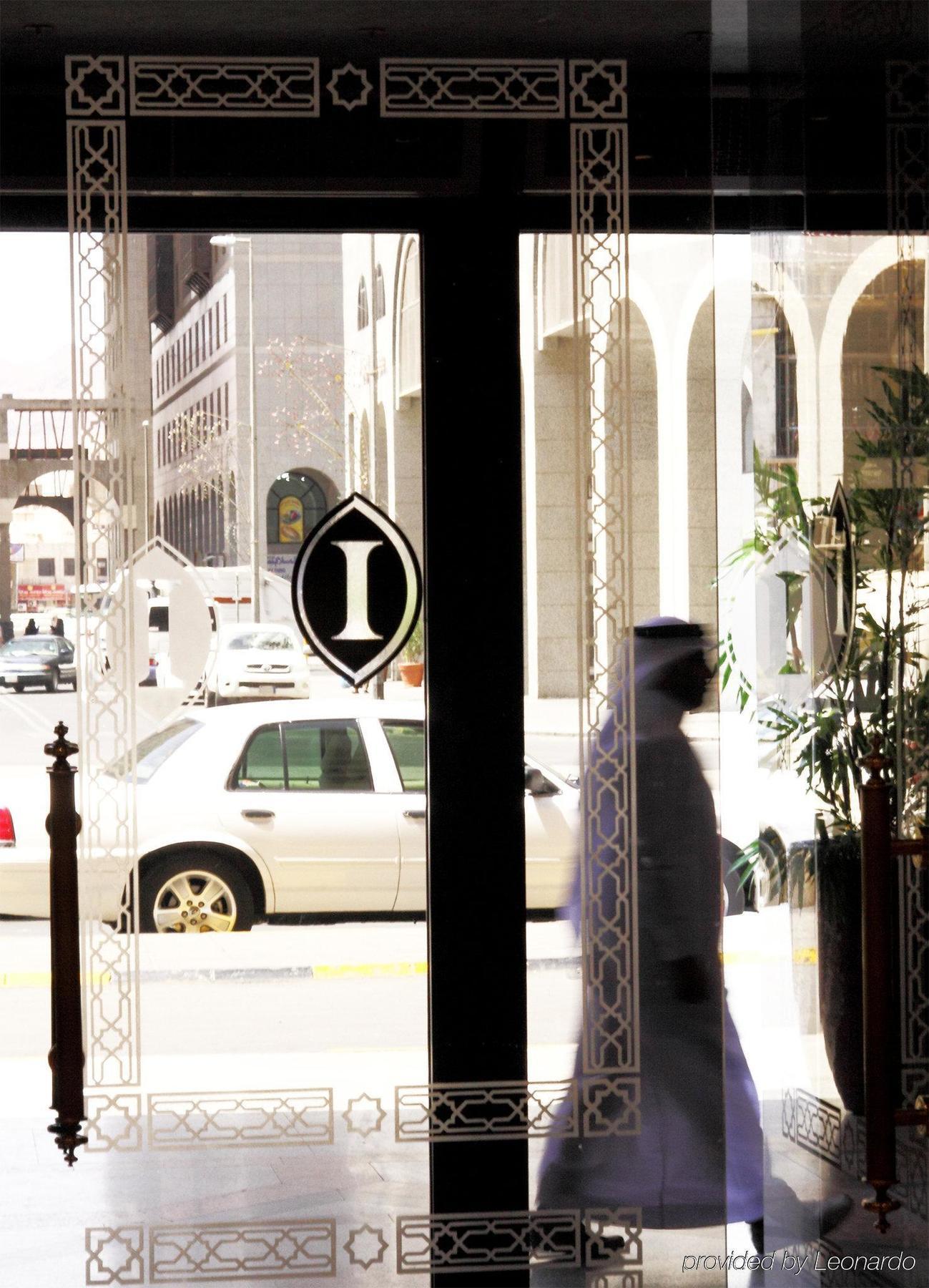 다르 알 이만 인터컨티넨탈 호텔 메디나 내부 사진 The photo shows a view through a glass door or window, featuring an elegant design with a prominent "I" symbol displayed. Through the glass, there are some parked vehicles visible, including a white car. In the foreground, a shadowy figure of a perso