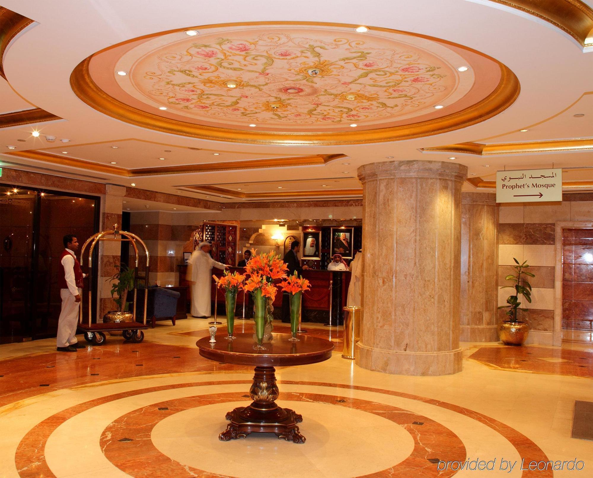 다르 알 이만 인터컨티넨탈 호텔 메디나 내부 사진 The photo depicts a luxurious hotel lobby. The ceiling features an ornate design with floral patterns, adding elegance to the space. In the center of the lobby, there is a decorative table with a bowl or vase of bright orange flowers. The flooring is