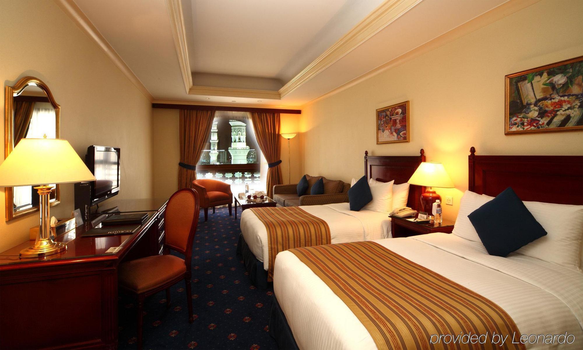 다르 알 이만 인터컨티넨탈 호텔 메디나 객실 사진 The photo shows a hotel room featuring two beds with white linens and brown striped throws. There is a wooden desk and chair, alongside a television. The walls are painted in a warm tone, and there are decorative paintings. Large windows with curtain