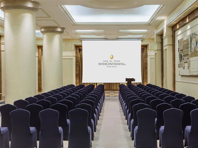 다르 알 이만 인터컨티넨탈 호텔 메디나 외부 사진 The photo shows a conference or meeting room set up. There are rows of dark blue chairs arranged facing a large screen at the front of the room. The screen displays the logo of the Intercontinental hotel, indicating that this space is likely intended