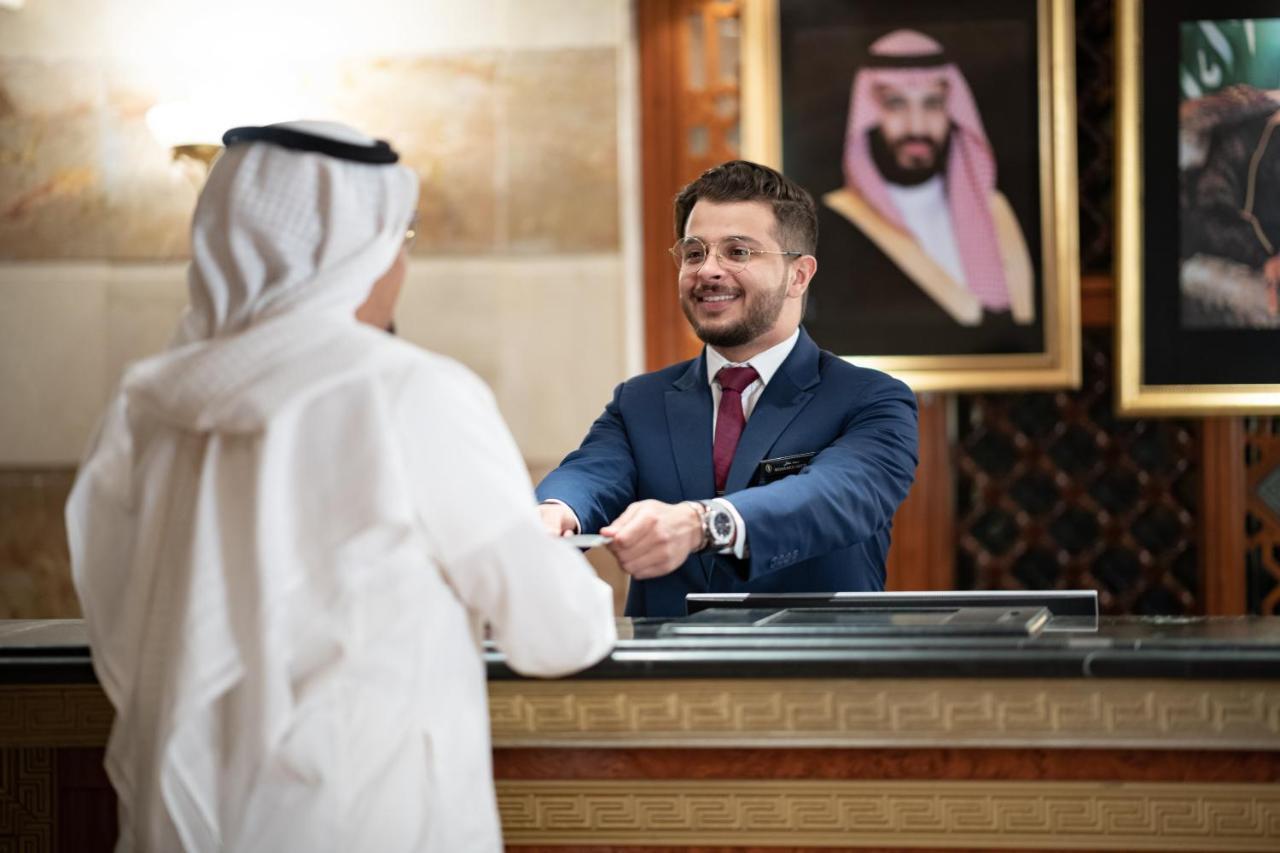 다르 알 이만 인터컨티넨탈 호텔 메디나 외부 사진 The photo shows a hotel reception area. A male receptionist, dressed in a navy suit with a tie, is smiling and handing an item to a guest who is wearing traditional white attire, possibly a kandura or thobe. The background features elegantly decorate